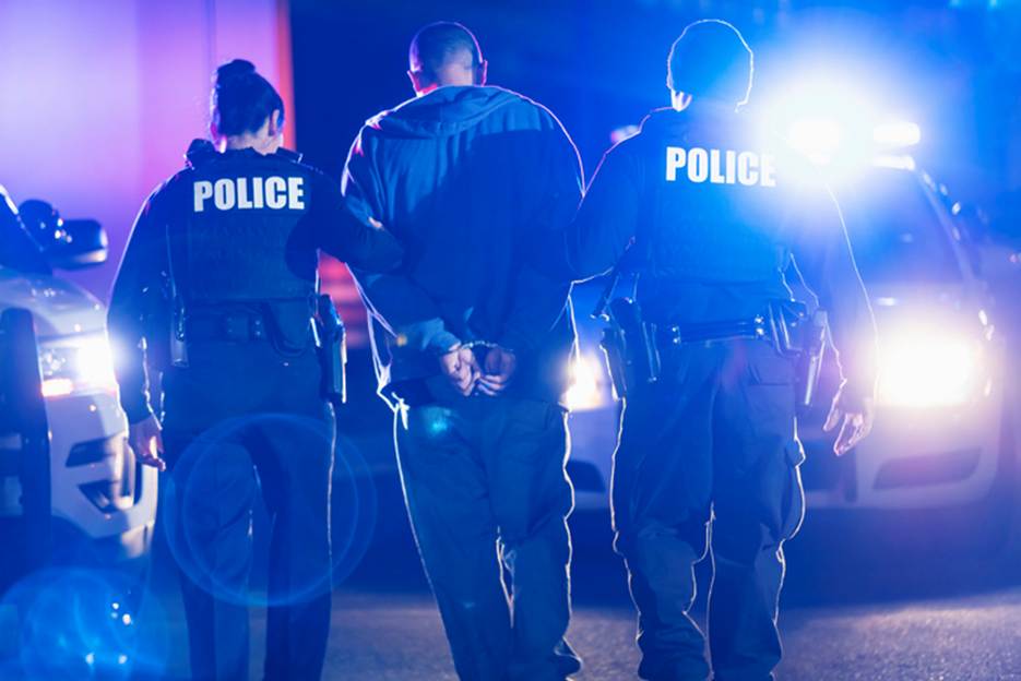 Two officers are arresting a suspect, whose hands are cuffed behind his back, with police cruisers behind them. The back of the officers' vests read "POLICE."